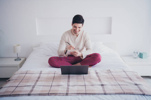 istock Photo of cheerful nice man sitting on bed writing notes weekly plan white light room indoors 2155861065
