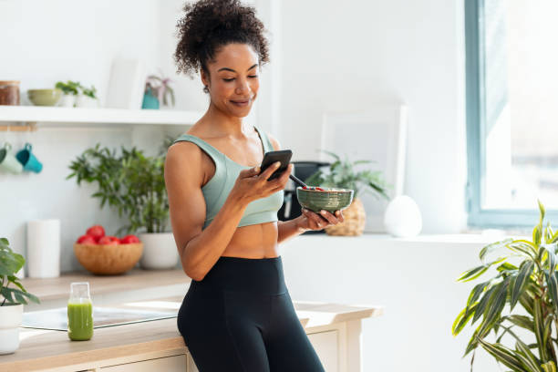 Athletic woman eating a healthy fruit bowl  while using smartphone in the kitchen at home
