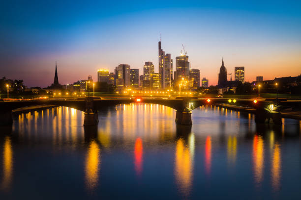 Frankfurt skyline at sunset with illuminated bridge and city lights reflecting on the river. Captures urban beauty and tranquility.