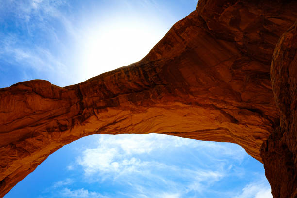 A striking image of a sunlit natural arch rock formation set against a bright blue sky with wispy clouds. The warm, reddish hues of the arch contrast beautifully with the cool tones of the sky, highlighting the dramatic natural beauty of the American Southwest desert. This photo captures the essence of a road trip adventure, showcasing the unique geological features and the breathtaking landscapes found along America's scenic highways.