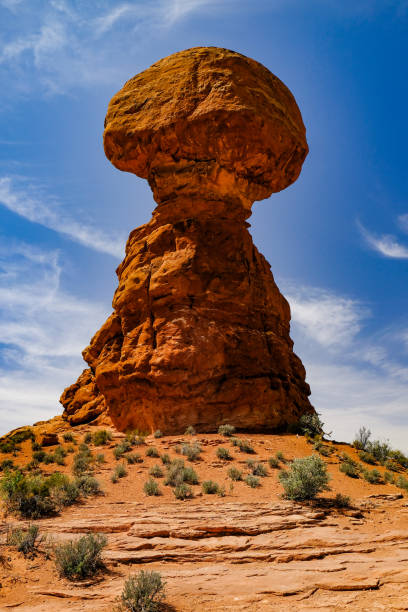 A towering balanced rock formation stands proudly against a vibrant blue sky in the American Southwest desert. The unique geological structure, with its large boulder precariously perched atop a narrow base, is surrounded by arid terrain and sparse vegetation. This image highlights the awe-inspiring natural wonders that can be discovered on road trips through the USA, capturing the essence of adventure and the rugged beauty of America's diverse landscapes.