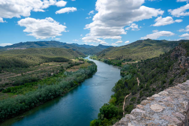 The Ebro river flows through a valley with mountains in the background. The water is calm and clear, and the sky is blue with some clouds. The scene is peaceful and serene