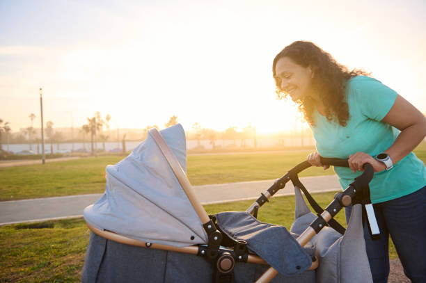 istock Side portrait of authentic happy mother looking and pushing infant baby stroller while walking in park at sunset. The concept of healthy active lifestyle. Daily outdoors walking and maternity leave 2155918545
