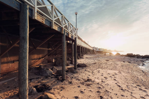 A tranquil beach scene beneath a wooden pier at sunset. The warm, golden light of the setting sun illuminates the sandy shore and rocky landscape, creating a peaceful and picturesque coastal view. Ideal for projects related to travel, coastal living, nature, and relaxation.