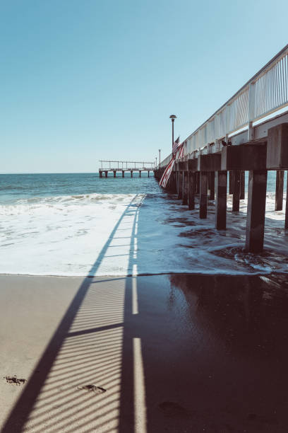 Beach scene with a pier extending into the calm ocean waters. The shadows cast by the pier's railing create interesting patterns on the sandy shore, while the gentle waves lap against the beach. Ideal for projects related to travel, coastal living, nature, and relaxation.