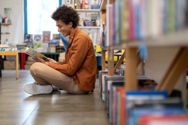 Black student guy reading book while sitting on floor near bookshelves in the university library, serene scene of immersive and traditional learning