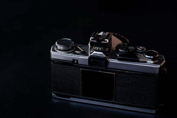 A vintage film camera with a black body and silver details, placed on a dark surface. The camera is shown from the back, highlighting its classic design and mechanical features.