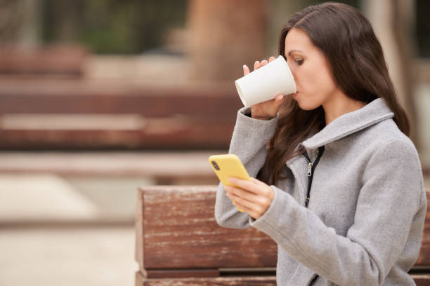 Female influencer sitting in a park drinking coffee and checking emails on smartphone