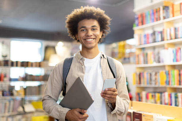 istock Happy black male student in library, holding digital tablet and cellphone, combining modern technology with traditional study methods 2155954001