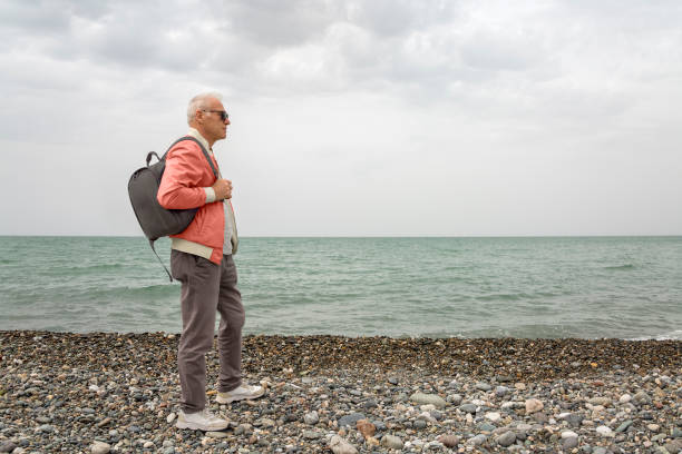 An elderly stylish man in a red windbreaker with a backpack on his shoulder stands on a pebble beach on a cloudy day. Full length