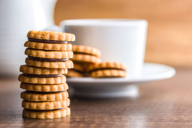 A close-up image of a stack of chocolate-filled cookies resting on a wooden table. A white cup and pitcher are visible in the background, out of focus. The cookies are golden brown in color, with a visible chocolate filling between each layer.
