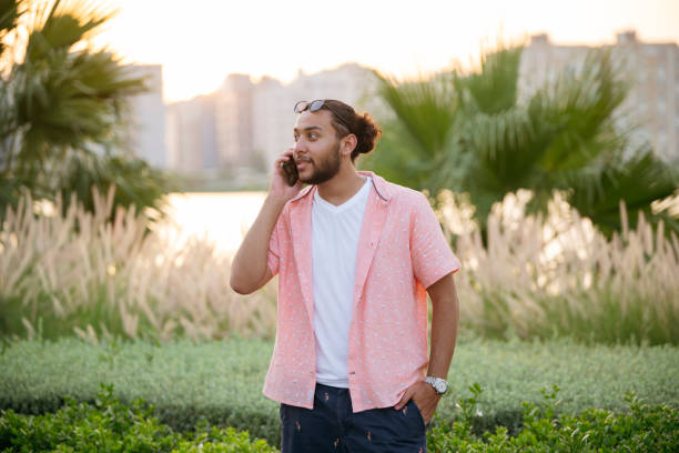 Generation Z Saudi man talking in lush public park in Jeddah. Man talking on phone in park part of sustainable urban greening project for vision 2030 arabian sunglasses stock pictures, royalty-free photos & images