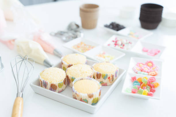 istock Selective focus on fresh baked cupcakes. Piping bags with pastel butter cream, icing candy, chocolate flakes or coated sugar and jelly in tray for decorating. Whisk, measuring spoons on white table 2156023991
