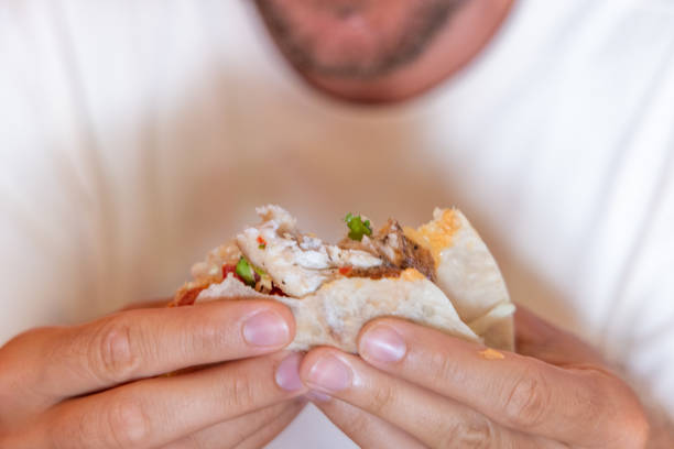 A detailed close-up shot of a person holding a taco filled with fresh ingredients, including shredded meat, vegetables, and cheese, wrapped in a soft tortilla. The person, wearing a white shirt, is about to take a bite, highlighting the delicious and appetizing nature of the meal.