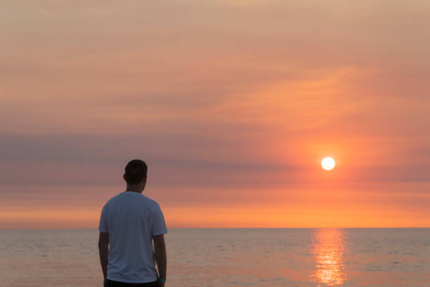 istock Man standing on beach sand looking towards horizon at orange sunset. Vacation concept. 2156040133