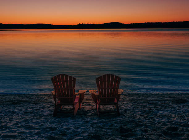 Two beach chairs sit empty on the shore at sunset by the water
