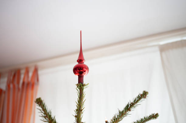 Decorated Christmas tree, real Nordmann fir in front of blurred room with orange curtains. A decorative red tree top. Unusual perspective.