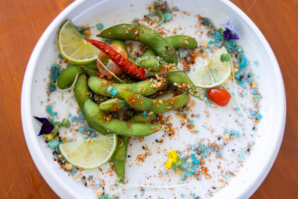 Close-up of a gourmet edamame salad garnished with lime slices, dried chili, colorful spices, and herbs on a white plate.