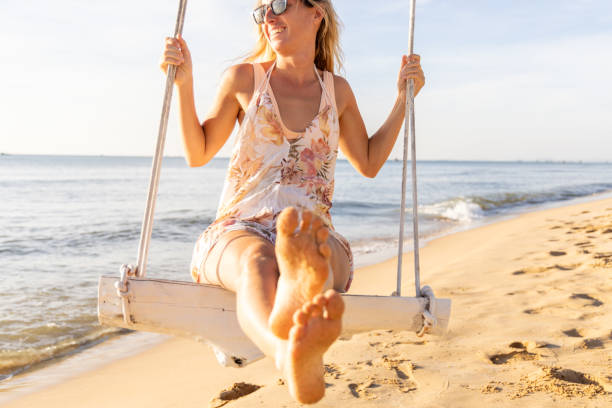 Girl reading book on wooden swing in mountains. Young girl lounges barefoot on a large wooden swing, peacefully reading a book with mountain scenery and blue sky behind her