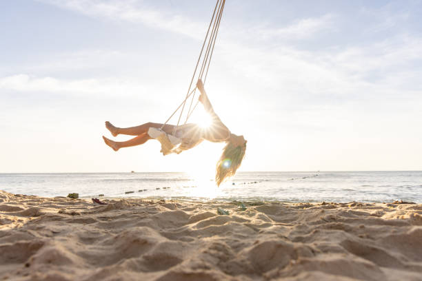 A woman in a floral dress swinging high above the sandy beach as the sun sets behind her. Her silhouette is highlighted by the warm, golden light of the setting sun, creating a magical and carefree atmosphere.