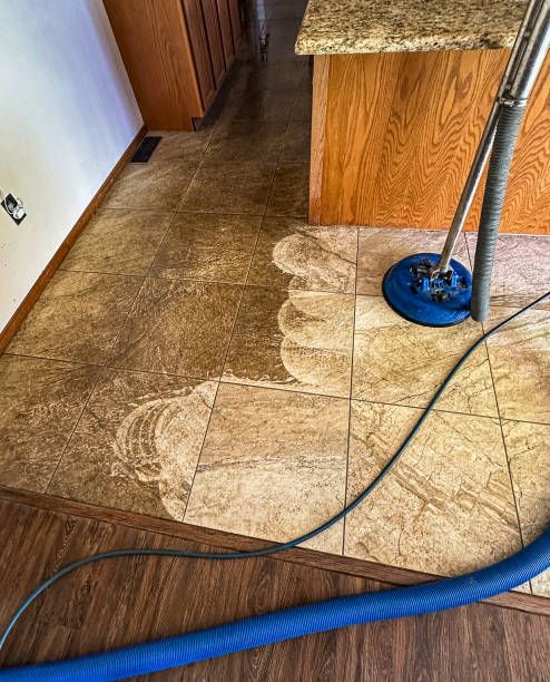 A professional floor cleaning machine in action, effectively removing dirt and grime from a tiled kitchen floor. The difference between the cleaned and uncleaned sections is clearly visible, showcasing the machine's powerful cleaning capabilities. The scene features part of a kitchen with wooden cabinetry and granite countertops. This image highlights the importance of regular deep cleaning for maintaining the appearance and hygiene of tile floors, making it ideal for promoting professional cleaning services and equipment.