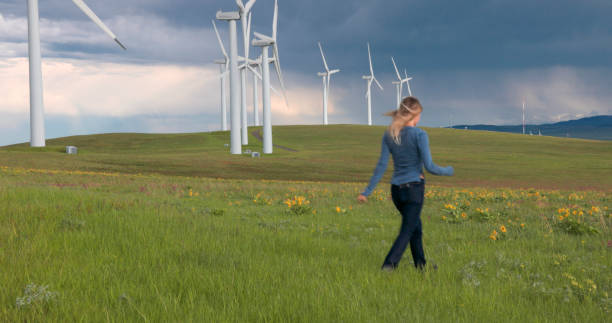 Woman in green shirt and linen trousers holding large rainbow pinwheel in wheat field with wind turbines. Concept of colorful protest, freedom and support for clean energy.