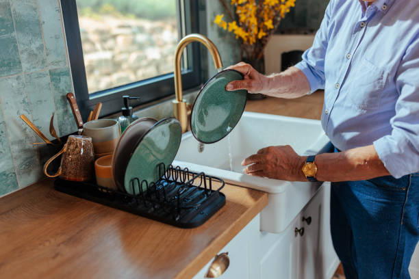 A senior white man standing next to a kitchen sink is putting a plate to be dried off after washing it.