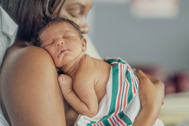 A new Mother, of African decent, holds her baby on her shoulder as she sits up in her hospital bed and they spend time bonding skin-to-skin.  The Mother has her eyes closed as she rests from exhaustion after childbirth.