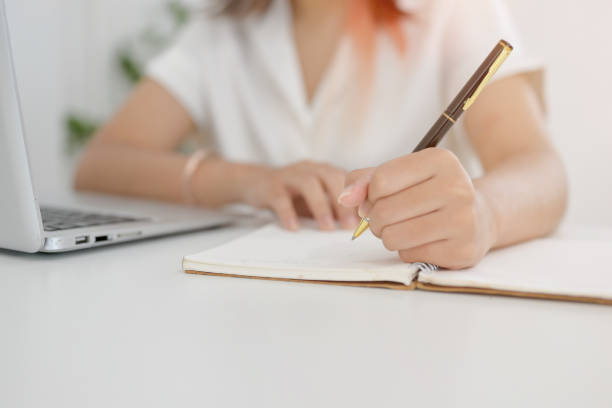 istock A cropped image of an Asian woman holding a pen, writing or taking notes in a notebook. 2156128369