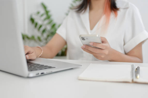 istock A cropped image of an Asian woman holding her smartphone while working on her laptop computer. 2156128385