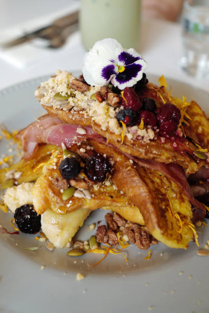 A delightful serving of crispy bacon strips stacked over golden brown toast on a blue plate. Accompanied by a small metal container of syrup and a sprig of rosemary, all beautifully arranged on a rustic wooden table. Perfectly captures the essence of a hearty morning meal.