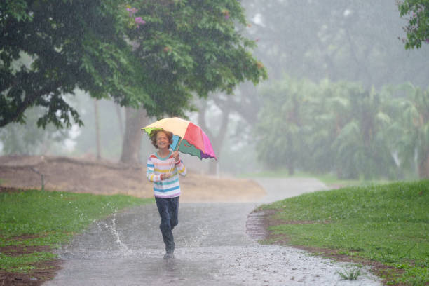 Child playing in autumn rain. Kid jumping in muddy puddle in beautiful park. Little boy with colorful umbrella under heavy fall shower. Stormy rainy weather fun.