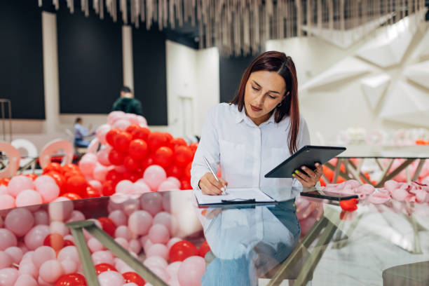 A decorator sits at a table in a restaurant and uses a digital tablet that displays detailed plans for the decoration and organization of the party, which she writes down