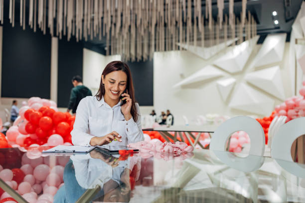 Smiling decorator sits at the table in the restaurant and talks on the phone with the client, writes down a detailed plan for the decoration and organization of the party