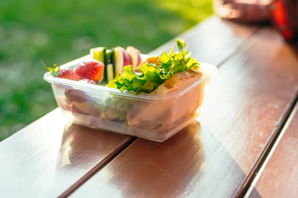 Plastic container with sandwich, vegetables and fruits on a table in a park in sunset