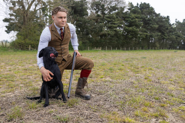 Medium shot of a young man crouched on one knee on a farmer's field. He is holding a cocked shotgun as he is going hunting. He has one arm around his hunting dog that is sitting next to him. The man is wearing traditional English hunting clothing.Videos are available similar to this scenario.