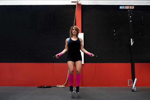Full length portrait of muscular woman in pink wristwraps and knee socks jumping rope in gym gym.
