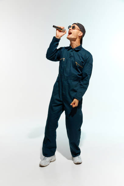 Artistic young man, musician dressed overalls with sunglasses singing with microphone against white studio background. Concept of music, festivals, contemporary art, hobby, creativity. Ad