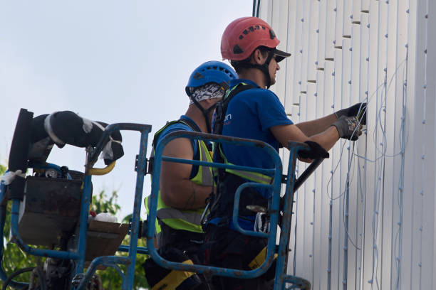 A low-angle shot captures two workers scaling a towering red electrical transmission tower against a pale, overcast sky. The tower's intricate steel lattice structure forms a stark geometric pattern, contrasting with the soft, diffused light. The workers, clad in safety harnesses and work gear, appear as small figures against the massive framework, emphasizing the scale of the structure and the height at which they are working. Thin electrical wires stretch horizontally across the frame, adding to the industrial aesthetic. The image highlights the challenging and potentially dangerous work of maintaining essential infrastructure.