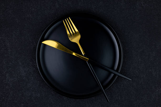 istock Black plate with fork and knife on black concrete background top view 2156202021