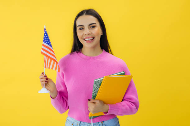istock Optimistic woman holding books in hand and American flag while standing in studio with yellow background supporting national holiday and feeling excited. 2156203974