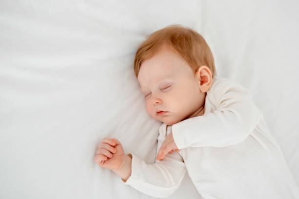 istock Baby boy sleeping under a blanket in a white bodysuit on the bed with his hand folded under his cheek, sweet healthy baby sleep at home 2156211226