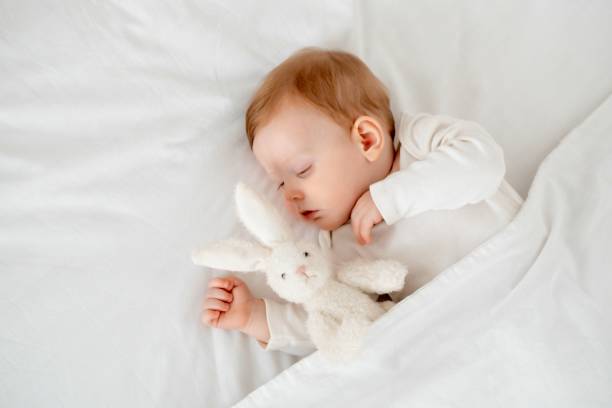 istock A baby boy sleeps under a blanket in a white bodysuit on the bed with his hand folded under his cheek and holding a soft toy, a sweet healthy baby sleep at home 2156211238