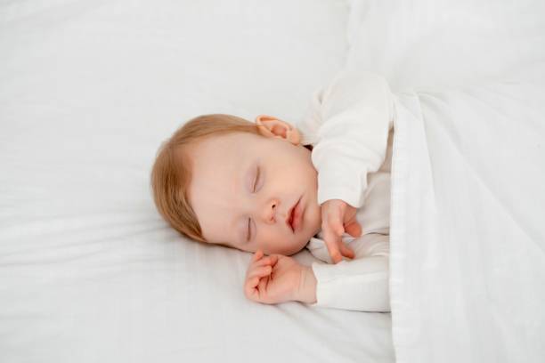 istock Baby boy sleeping under a blanket in a white bodysuit on the bed with his hand folded under his cheek, sweet healthy baby sleep at home 2156211251