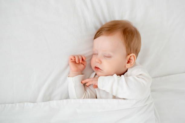 istock Baby boy sleeping under a blanket in a white bodysuit on the bed with his hand folded under his cheek, sweet healthy baby sleep at home 2156211259
