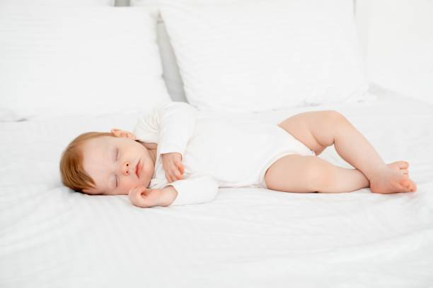istock Baby boy sleeping under a blanket in a white bodysuit on the bed with his hand folded under his cheek, sweet healthy baby sleep at home 2156211270