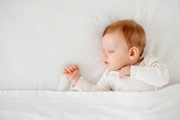 istock Baby boy sleeping under a blanket in a white bodysuit on the bed with his hand folded under his cheek, sweet healthy baby sleep at home 2156211280
