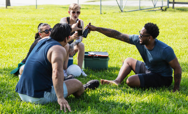A multiracial group of four friends hanging out together on a sunny day at the park, sitting in a circle on the grass, enjoying refreshments. The main focus is on the non-binary person sitting on the cooler and the Hispanic woman sitting next to her. They are both in their 20s. On the other cooler is a solar panel recharging a mobile phone.