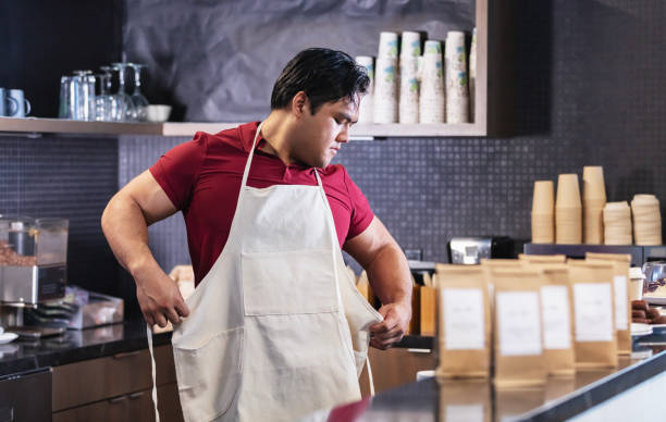 A young Asian man working behind the counter in a coffee shop, putting on an apron. This responsible small business uses compostable cups, utensils and brown paper bags for merchandise.