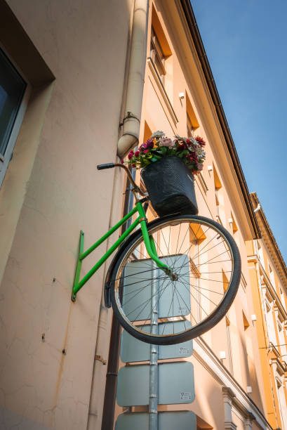 Creative Urban Bicycle Installation in Riga, Whimsical and Vibrant Urban installation with a green bicycle on a wall, adorned with colorful flowers above gray electrical boxes. Possibly located in old town Riga, under a clear sky. unusual basket stock pictures, royalty-free photos & images